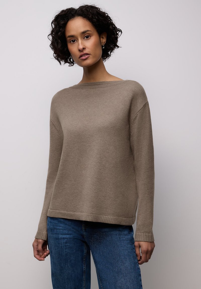Femme aux cheveux bouclés et foncés portant un pull en tricot beige et un jean bleu, debout devant un fond gris clair uni.