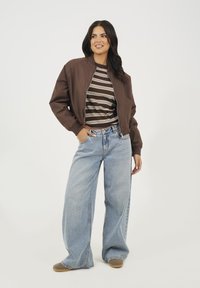 Brązowa kurtka bomberka na paski, zestawiona z jasnoniebieskimi szerokimi spodniami i szarymi butami wsuwanymi. Prosty, casualowy strój o luźnym kroju.