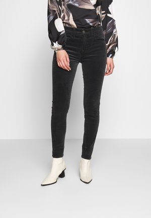Femme portant un jean skinny noir, des bottines blanches et un chemisier à manches longues avec un motif de feuilles marron et beige, debout devant un fond uni.