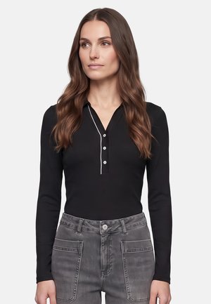 Woman with long wavy brown hair wears a black long-sleeve top with white trim and gray high-waist jeans, looking slightly right.