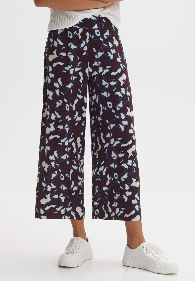 Vue en longueur des jambes d'une personne portant un pantalon large et court avec un motif floral abstrait bleu foncé et blanc, ainsi que des baskets blanches.