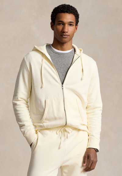 Camisola com capuz creme com fecho de correr, bolsos frontais e textura leve, usada sobre uma camisola cinzenta de gola redonda, combinada com calças a condizer.