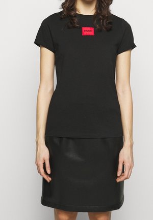 Schwarzes Baumwoll-T-Shirt mit kurzen Ärmeln, das mit einem roten rechteckigen Logo-Patch versehen ist. Getragen mit einem schwarzen Kunstlederrock.