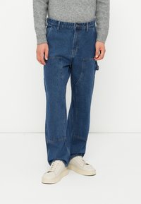 Zilā denim auduma cargo bikses ar brīvu piegriezumu, vairākām kabatām un redzamām dūriena detaļām, apvienotas ar krēma krāsas sporta apaviem.