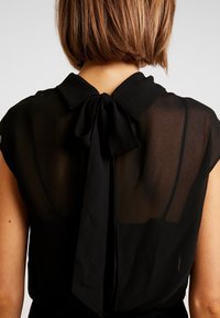 Musta lyhythihainen paita, jossa on läpikuultava selkä, kaulus ja suuri solmukohta kaulassa, pehmeä materiaali ja vartalonmyötäinen muotoilu.