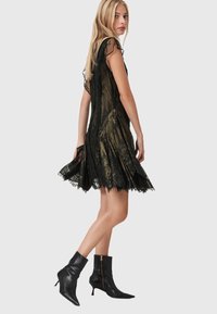 Donna bionda che indossa un abito da cocktail in pizzo nero e stivali alla caviglia neri a punta, in posa di lato contro uno sfondo semplice.