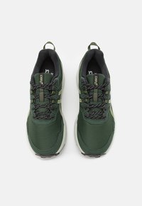 Zapatillas deportivas verdes con parte superior de malla, acentos en negro, suela texturizada y costuras en contraste. Presenta cordones y una tira de agarre en el talón.