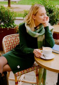 Rochie tricotată verde cu un tiv cu nervuri, asortată cu o eșarfă în carouri albă și verde deschis. O cană albă pe o masă aurie și o carte albastră aproape.