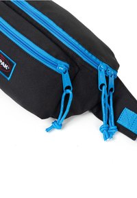 Černá látková ledvinka se modrými zipy a detaily. Obsahuje dvě zipové přihrádky, odolný materiál a praktický nastavovací popruh.