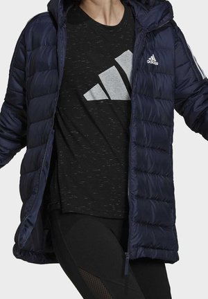 Personne portant une doudoune Adidas bleu marine par-dessus un t-shirt noir Adidas et un legging noir, debout les bras légèrement levés.