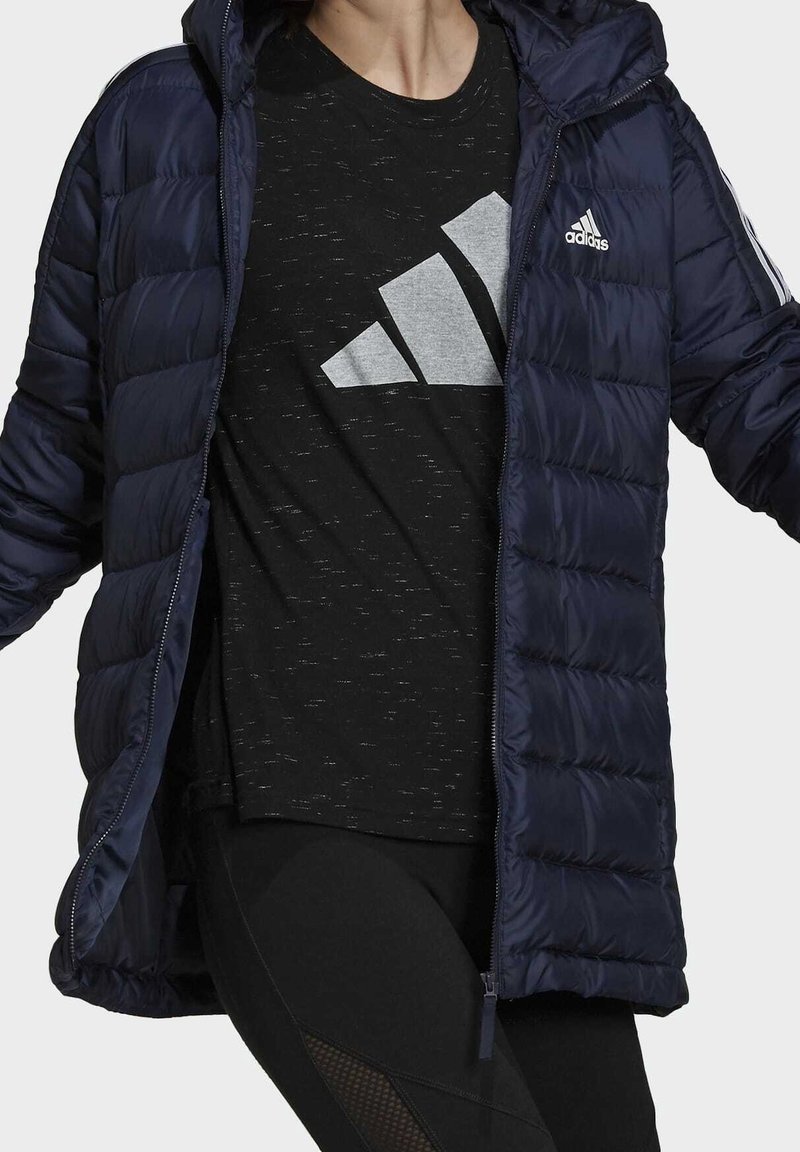 Personne portant une doudoune Adidas bleu marine par-dessus un t-shirt noir Adidas et un legging noir, debout les bras légèrement levés.