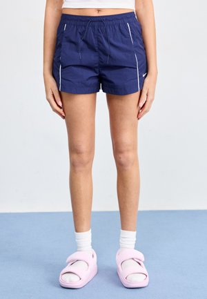 Marineblauwe shorts met een elastische tailleband en witte zijbuis, gecombineerd met roze sandalen en witte enkelsokken.