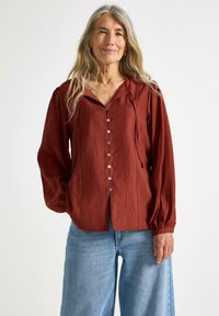 Maronfarvet bluse med krave, frontlukning med knapper og lange, voluminøse ærmer. Tekstureret stof og afslappet pasform i kombination med blå denimjeans.