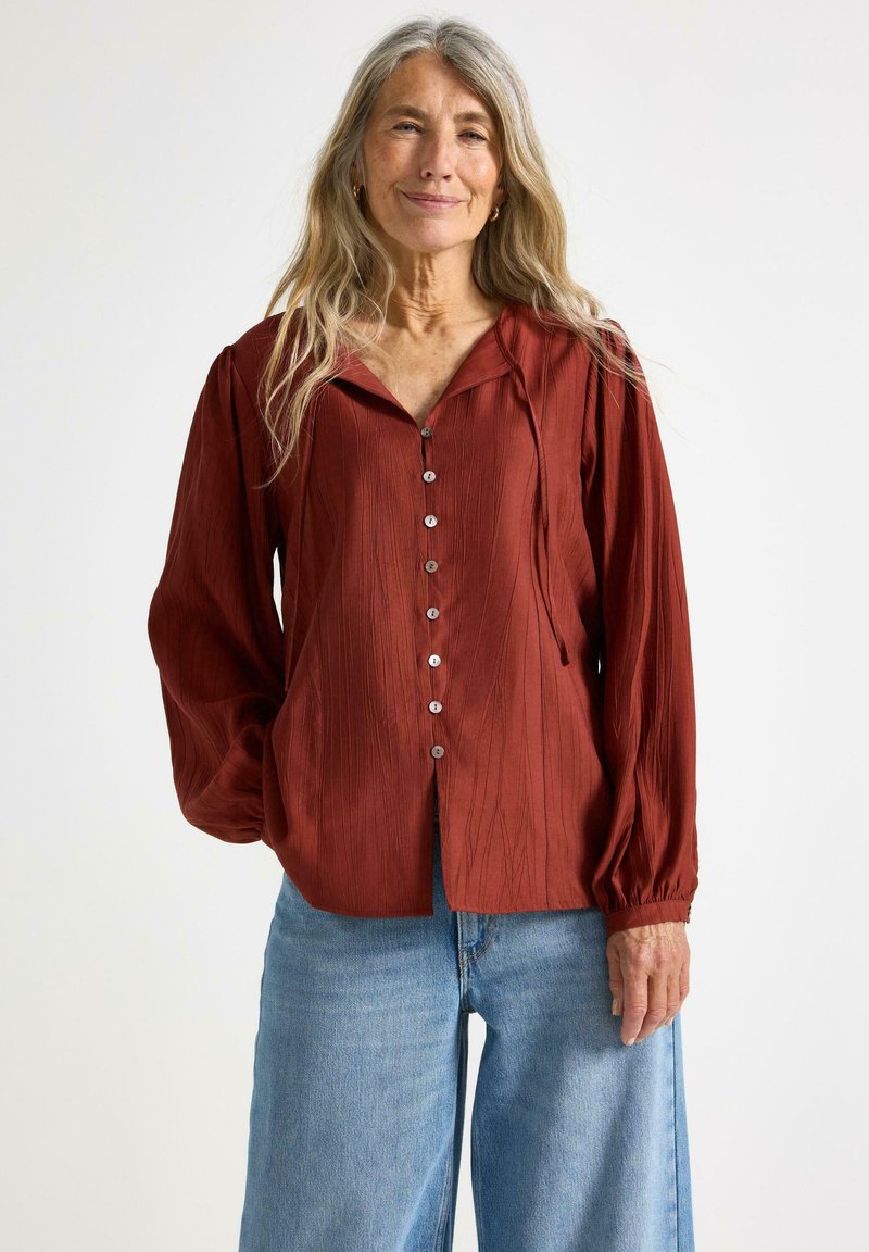 Maronfarvet bluse med krave, frontlukning med knapper og lange, voluminøse ærmer. Tekstureret stof og afslappet pasform i kombination med blå denimjeans.