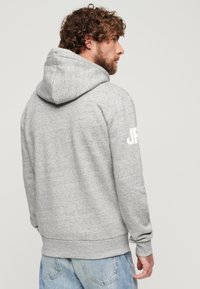 Felpa grigia con una texture morbida, dotata di cappuccio e logo bianco "JR" sulla manica destra; abbinata a jeans azzurri.