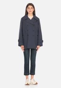 Cappotto impermeabile doppiopetto di colore navy con ampi revers, bottoni neri e tasche laterali. Indossato sopra una maglietta chiara e jeans scuri con risvolti.