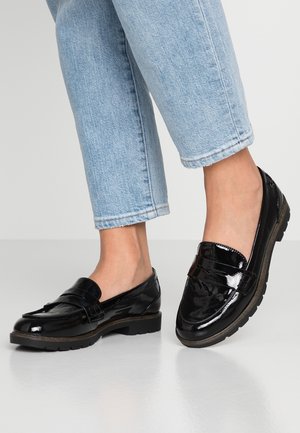 Fødder iført skinnende sorte loafers kombineret med lyseblå cowboybukser, stående på en reflekterende grå overflade.