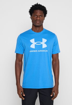 Man die een felblauw Under Armour T-shirt en zwarte broek draagt, staat tegen een effen lichtgrijze achtergrond.