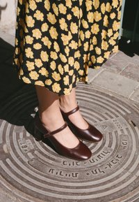 Hnědé dámské boty typu Mary Jane s kulatou špičkou a blokovým podpatkem, s páskem přes nárt. Umístěné na textuře kanálové poklopky.