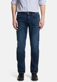 Jeans in denim blu scuro con taglio dritto, caratterizzati da cinque tasche e chiusura con zip. Indossati con una camicia a righe leggera e scarpe nere.