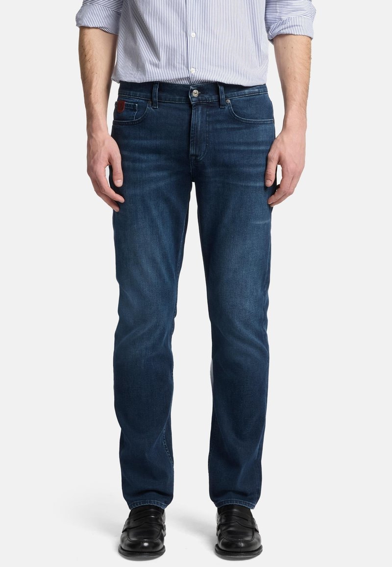 Jeans in denim blu scuro con taglio dritto, caratterizzati da cinque tasche e chiusura con zip. Indossati con una camicia a righe leggera e scarpe nere.