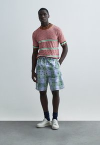 Μπλούζα πλεκτή με κόκκινο και λευκό σχέδιο, με πράσινες και μαύρες ρίγες, συνδυασμένη με μπλε και λευκά καρό σορτς και λευκά αθλητικά παπούτσια.