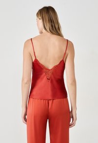 Czerwony satynowy top na ramiączkach z regulowanymi paskami oraz koronkowym akcentem na plecach w kształcie litery V. Dopasowane satynowe spodnie dopełniają całość.