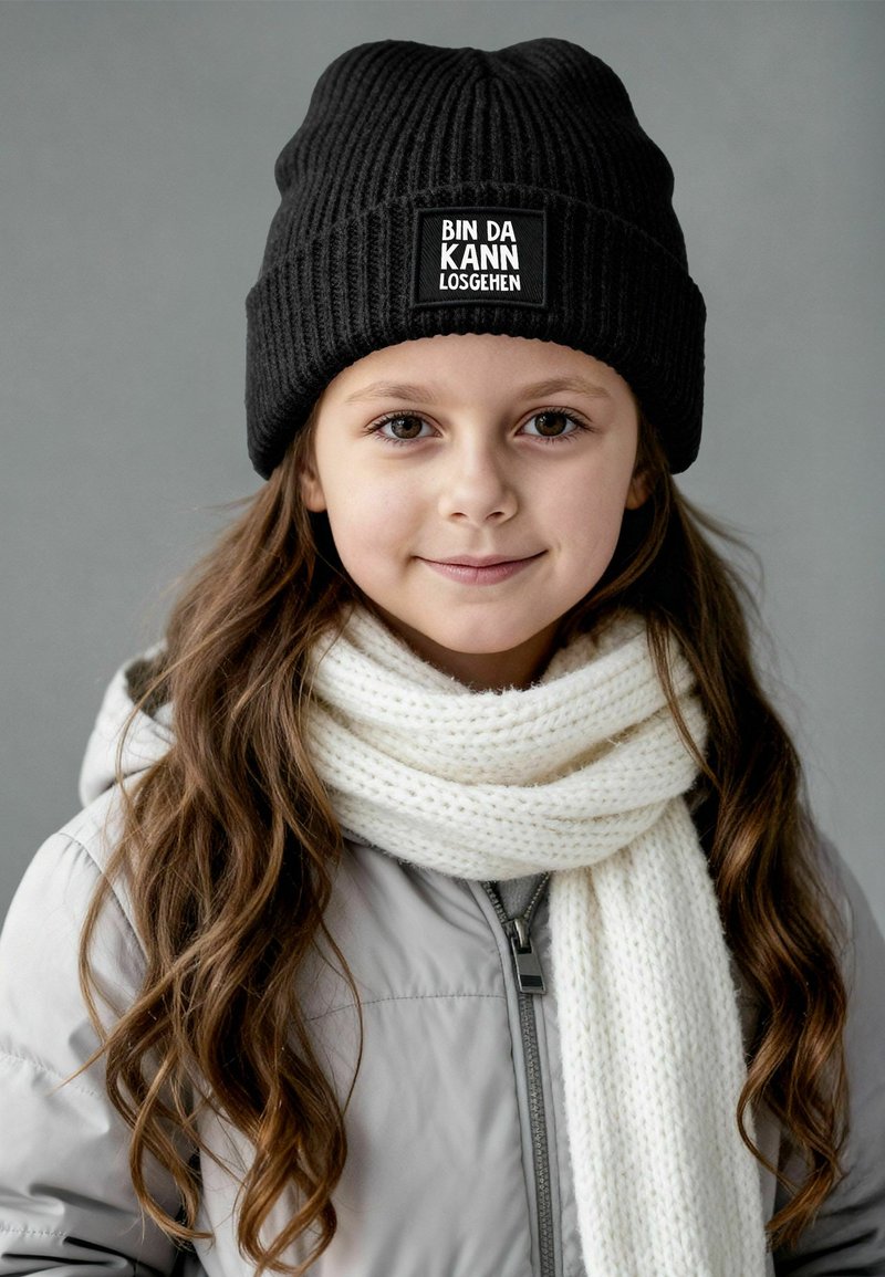 Black ribbed beanie with a square patch reading "BIN DA KANN LOSGEHEN," paired with a white knitted scarf and a light gray puffer jacket.