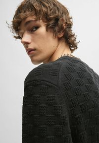 Schwarzer Strickpullover mit strukturiertem Karomuster, Rundhalsausschnitt und leicht tailliertem Design, mit sichtbaren Nähdetails.