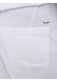 Tejido de camisa de lino blanco con un bolsillo en el pecho izquierdo que incluye una pequeña etiqueta que dice "JACK & JONES EST. 1990." La textura parece transpirable y ligera.