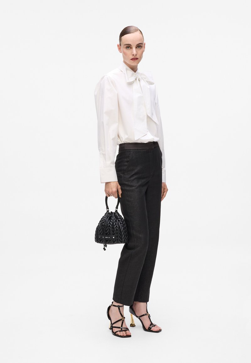 Vrouw in een witte blouse met strik, zwarte getailleerde broek, zwarte hoge sandalen en met een kleine zwarte, gequiltte handtas.