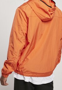 Orange letvægtsjakke med hætte, elastiske manchetter og en let skinnende tekstur. Har en afslappet pasform og et simpelt design.