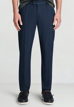 Homme portant un pantalon de costume bleu marine et des chaussures de ville noires à lacets, debout devant un fond clair uni.