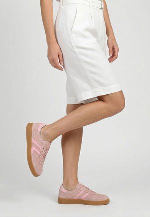 Frau trägt weiße knielange Shorts und hellrosa Sneaker mit Gummisohlen, steht mit einem Bein nach hinten gebogen vor einem einfarbigen Hintergrund.