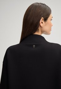 Zwarte sweatshirt met een hoge kraag, gladde textuur en een prominent rond logo op het achterste nekgebied. Eenvoudig, modern ontwerp.