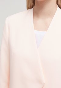 Jasnoróżowa marynarka o miękkiej fakturze, z kołnierzem szalowym i luźnym krojem, noszona na białej bluzce.