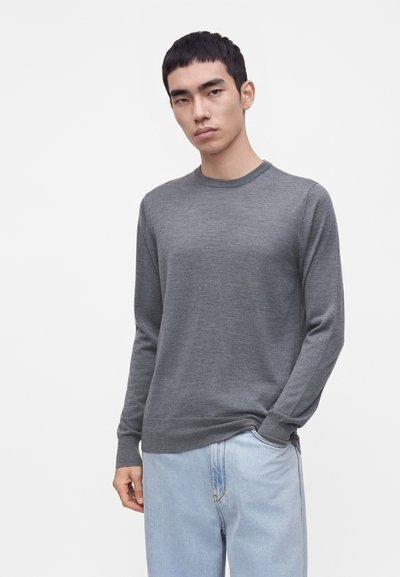 Jeune homme aux cheveux courts et noirs portant un pull gris à manches longues et un jean bleu clair, debout avec une main dans la poche.