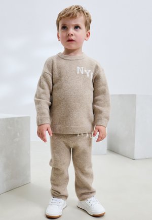 Batoľa s krátkymi blond vlasmi, oblečené v béžovom pletenom svetri a nohaviciach s bielymi teniskami, stojace v minimalisticky svetlej miestnosti.