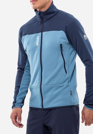 Homme portant une veste polaire zippée bicolore bleue avec des poches zippées latérales et un logo sur la poitrine et la manche.