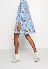 Person klädd i en blå blommönstrad klänning och vita högklackade ankelskor, går mot en helt vit bakgrund.