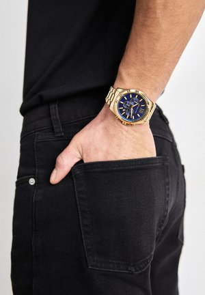 Mano izquierda con reloj de oro y esfera azul colocada dentro del bolsillo trasero de unos vaqueros negros, persona con camisa negra.
