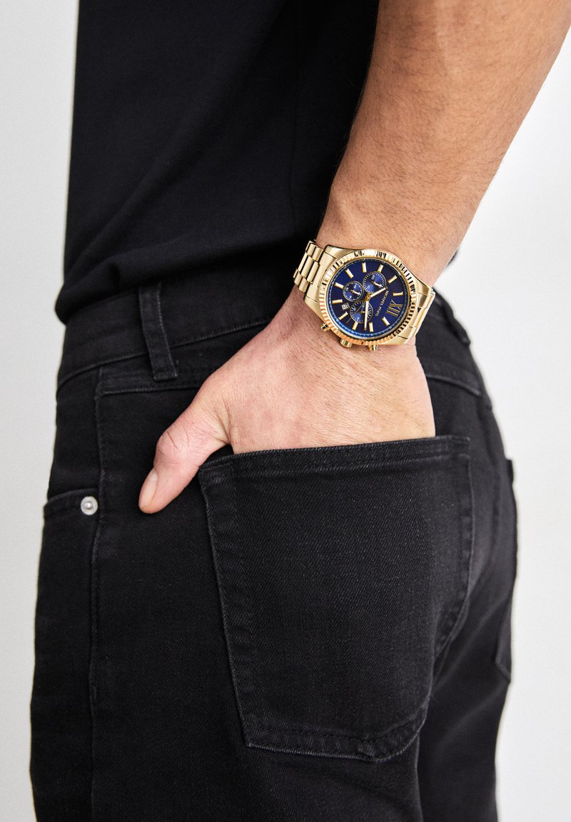 Linke Hand mit goldener Uhr und blauem Zifferblatt in der Gesäßtasche einer schwarzen Jeans, Person trägt schwarzes Hemd.