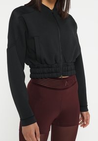Frau in einer schwarzen, kurz geschnittenen Zip-Jacke und hoch taillierten, kastanienbraunen Sportleggings mit einem Netzpaneel.