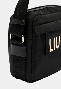 Borsa a tracolla nera in nylon con cinturino testurizzato, chiusura a zip e un logo ben visibile di colore chiaro sul frontale. Dettagli in hardware dorato.