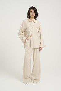 Chemise beige surdimensionnée avec poches avant, associée à un pantalon large. Texture lisse avec une silhouette décontractée et fermeture à boutons.