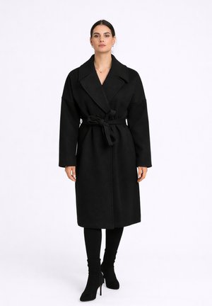 Mujer con un abrigo largo negro con cinturón, leggings negros y botines negros de tacón, de pie contra un fondo blanco.