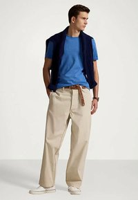 Homme debout les mains dans les poches, portant un t-shirt bleu, un pantalon large beige avec une ceinture tressée, des chaussures blanches et un pull bleu marine drapé sur les épaules.