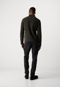 Suéter de canalé verde oscuro con un alto cuello, combinado con pantalones negros y zapatos marrones, visto desde atrás.
