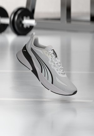 Λευκό και μαύρο αθλητικό παπούτσι Puma που αιωρείται σε ένα γυμναστήριο με θολό εξοπλισμό άρσης βαρών στο φόντο.