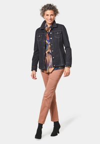 Schwarze Jeansjacke mit hellen Nähdetails, getragen über einem gemusterten Hemd, kombiniert mit pinken Hosen und schwarzen Stiefeletten.